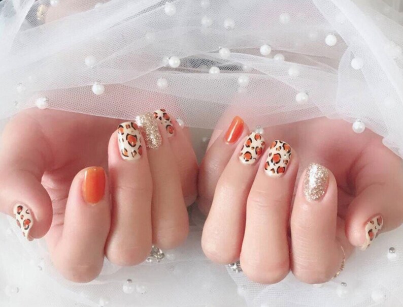 May include: Manicured nails with a variety of designs. Some nails are painted a solid orange, while others feature a leopard print pattern with orange spots on a cream background. One nail has a gold glitter finish.