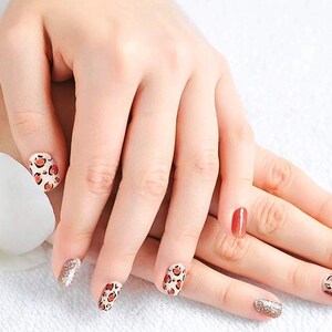 May include: Close-up of hands with manicured nails. Some nails are painted with a leopard print design, others are solid colors of red and orange, and some are glittery. A white flower is in the background.