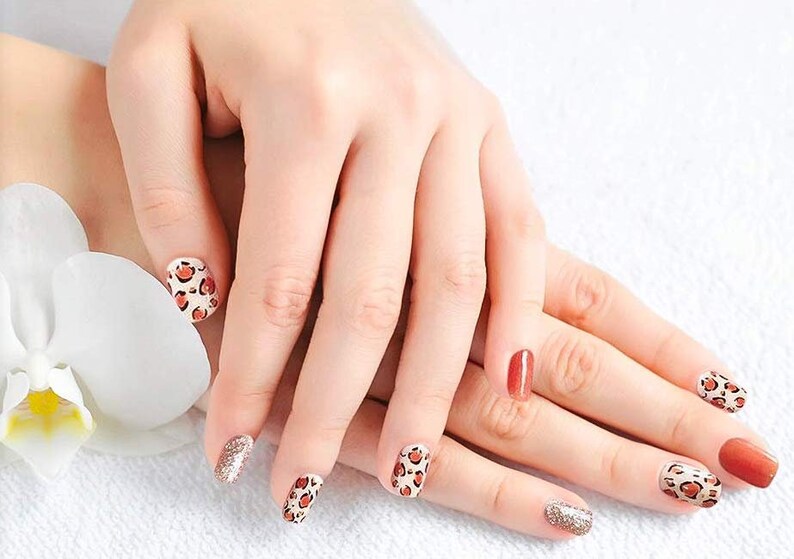 May include: Close-up of hands with manicured nails. Some nails are painted with a leopard print design, others are solid colors of red and orange, and some are glittery. A white flower is in the background.
