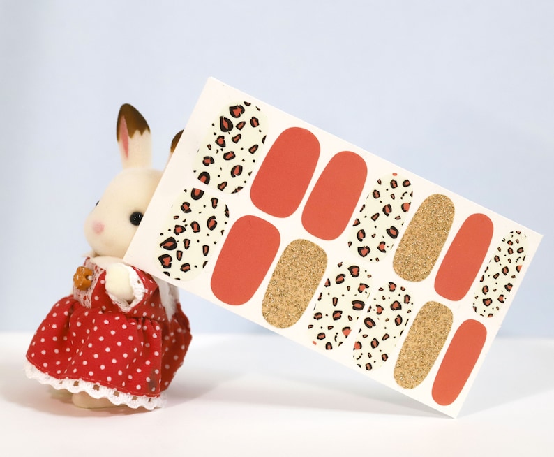 May include: Nail art stickers featuring a mix of designs. The sheet includes solid orange, gold glitter, and leopard print patterns in black and red. The stickers are displayed against a light blue background.