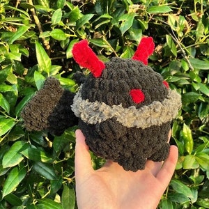 May include: A black plush toy with red horns and a grey fur trim around the neck. The toy is being held in a person's hand.
