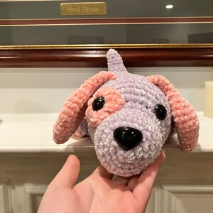 May include: A crocheted plush toy dog with a purple body and pink ears. The dog has black eyes and a black nose.