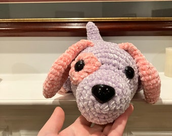 May include: A crocheted plush toy dog with a purple body and pink ears. The dog has black eyes and a black nose.
