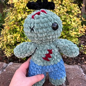 May include: A green crocheted plush toy zombie with a black crocheted hairpiece, red stitches, and blue crocheted shorts.