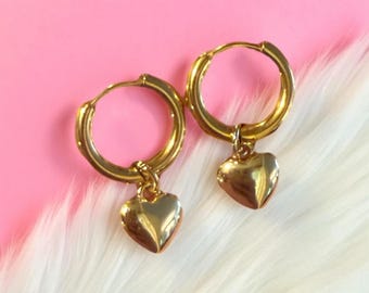 Puede incluir: Un par de pendientes de aro dorados con colgantes en forma de corazón. Los pendientes se muestran sobre una superficie de piel sintética blanca sobre un fondo rosa. Diseño sencillo y elegante.