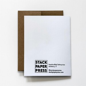 May include: A white rectangular greeting card with a brown kraft paper envelope. The back of the card features a black square logo with the text STACK PAPER PRESS, followed by the text handcrafted letterpress mchenry, il @stackpaperpress stackpaperpress.com.