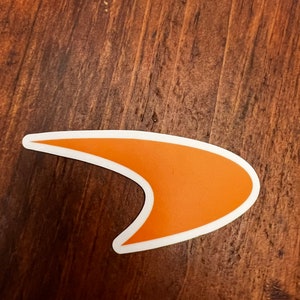 Puede incluir: Pegatina naranja en forma de boomerang con contorno blanco sobre una superficie de madera marrón.