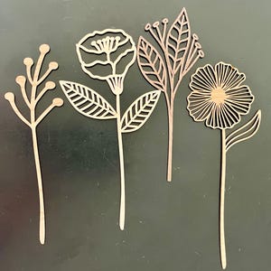 May include: Four wooden floral cutouts in various designs. The cutouts include a flower with a detailed center, a daisy-like flower, and two other botanical shapes. The wood has a natural, light brown color.