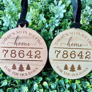 May include: Two round wooden ornaments with black ribbons. The ornaments have the text "THERE'S NO PLACE LIKE home 78642 FOR THE HOLIDAYS" engraved on them, along with three small Christmas trees. The ornaments are displayed on a bed of green foliage.