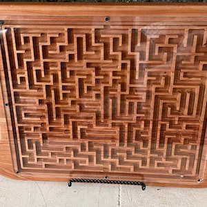May include: Wooden maze puzzle tray with a clear acrylic top. The tray is rectangular with rounded corners and built-in handles. The maze is carved into the wood, creating a complex path. The wood has a warm, reddish-brown tone.