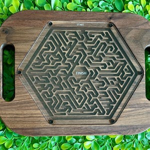 May include: A wooden tray with handles features a hexagonal maze game with a clear acrylic top. The maze has a start and finish point.