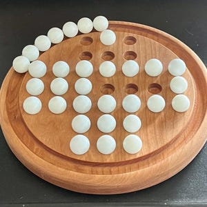 May include: A wooden solitaire board with a circular design and a series of drilled holes. The board is filled with white marbles, arranged in a pattern. The board is made of light brown wood.
