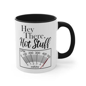 May include: White ceramic coffee mug with a black rim and handle. The mug features a black and white graphic of a gauge with the text "Hey There, Hot Stuff" and "CPM 0 100 200 300 400 500 mR/hr".