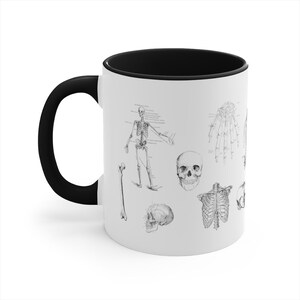 May include: White ceramic coffee mug with a black handle and black rim. The mug features a black and white anatomical illustration of a human skeleton, including a full skeleton, skull, rib cage, and bones.