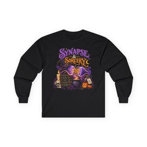 May include: A black long-sleeve shirt featuring a graphic print with the text "SYNAPSE & SORCERY". The design includes a purple witch hat, a stack of books, a skull, a lit candle, a bubbling cauldron, and orange pumpkins in a vibrant style.