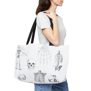 May include: White tote bag featuring detailed black and white anatomical illustrations of the human skeletal system. The bag has black shoulder straps and is carried over the shoulder.