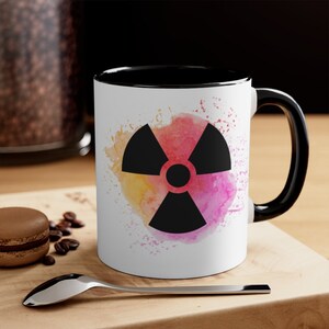 May include: White ceramic mug with a black rim and handle. The mug features a black radiation symbol on a watercolor background with pink, yellow, and orange hues.