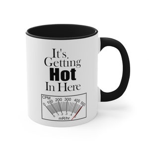 May include: White ceramic coffee mug with a black rim and handle. The mug has a black graphic that reads "It's Getting Hot In Here" with a black and white gauge underneath that reads "CPM 0 100 200 300 400 500 mR/hr".