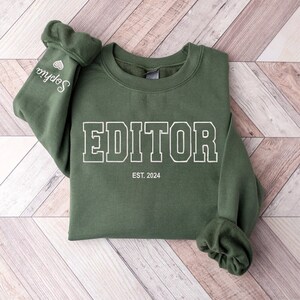 May include: A green sweatshirt with the word "EDITOR" printed in white with a black outline. The text "EST. 2024" is printed below the word "EDITOR".