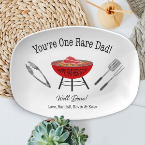 May include: White oval serving platter with a red and black grill, a pair of tongs, and a spatula and fork. The text on the platter reads "You're One Rare Dad!" and "Happy Father's Day". Below the grill is the text "Well Done! Love, Randall, Kevin & Kate".