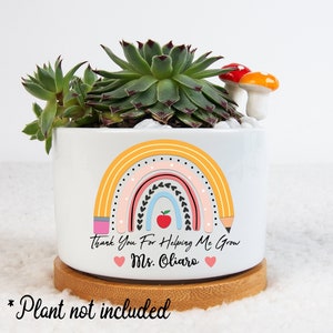 May include: White ceramic planter with a rainbow design, a red apple, and the text "Thank You For Helping Me Grow Ms. Oliaro." The planter holds a succulent and decorative mushrooms. It sits on a round wooden base.