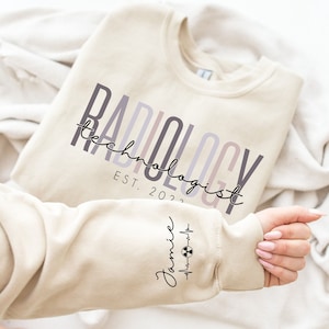 May include: A beige sweatshirt with the text "RADIOLOGY technologist EST. 2023" printed on the front. The left sleeve has a small graphic of a heart beat with a radiation symbol and the text "Jamie".