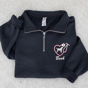 May include: A navy blue quarter-zip sweatshirt with a pink heart-shaped graphic featuring a dog silhouette, paw print, and stethoscope. The name "Bush" is printed below the graphic. The sweatshirt has a folded collar and long sleeves.