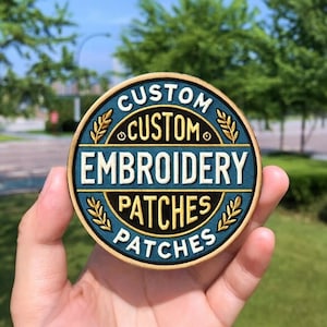 May include: A round, custom embroidery patch with a teal background and gold accents. The patch features the words "CUSTOM EMBROIDERY PATCHES" in a circular design. The patch is held in front of a blurred green background.