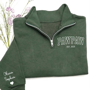 May include: A dark green quarter-zip sweatshirt with the word "PAWPAW" and "EST. 2025" printed in white. The sleeve has the names "Olivia Sophia" and a white heart. The sweatshirt has a silver zipper.