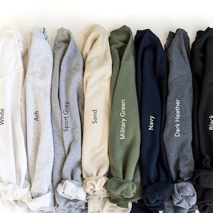 May include: A row of ten folded sweatshirts in various colors, including white, ash, sport grey, sand, military green, navy, dark heather, and black. The colors are labeled below each sweatshirt.