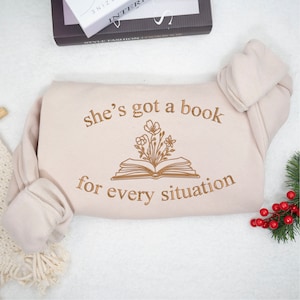 May include: A beige sweatshirt with the text "she's got a book for every situation" and a drawing of an open book with flowers growing out of it.
