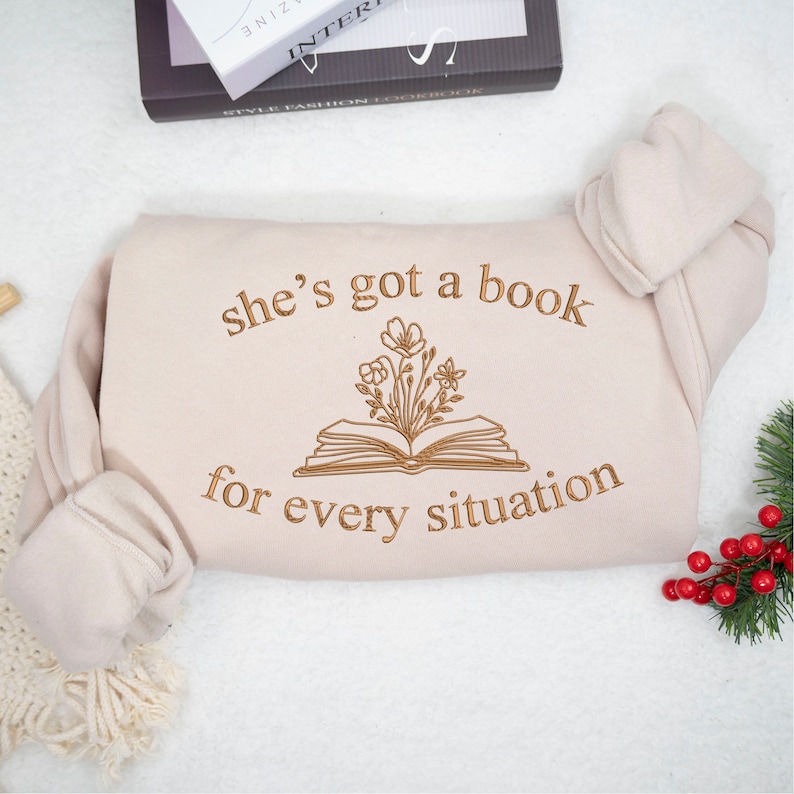 May include: A beige sweatshirt with the text "she's got a book for every situation" and a drawing of an open book with flowers growing out of it.