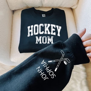 May include: Black sweatshirt with the text "HOCKEY MOM" printed on the front. The sleeve has the text "JOHN SOPHIA" and a graphic of two hockey sticks crossed over a puck.