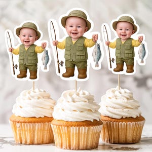 May include: Three cupcakes topped with white frosting. Each cupcake has a decorative topper featuring a cartoon baby dressed as a fisherman, holding a fishing rod and a fish. The baby wears a khaki hat, vest, and boots.