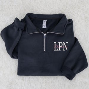 May include: A navy blue quarter-zip sweatshirt with the letters "LPN" in white and the name "Sherri" in pink script. The sweatshirt has a collar and a silver zipper. The sleeves are cuffed.