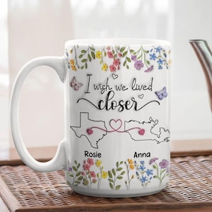 May include: White ceramic mug with a floral design and the text "I wish we lived closer." The mug features a map of Texas and another state, connected by a heart. Names "Rosie" and "Anna" are below the maps. Butterflies and flowers decorate the mug.