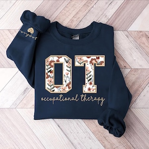 May include: A navy blue sweatshirt with the letters "OT" in white with a floral pattern. The letters are outlined in gold and the text "occupational therapy" is printed below.