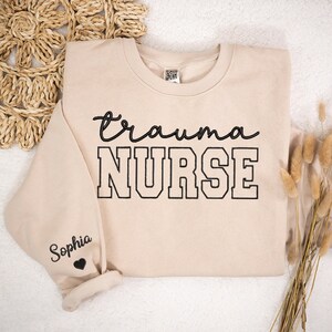 May include: A beige sweatshirt with the text "trauma NURSE" in black lettering. The text "Sophia" is embroidered on the left sleeve with a small black heart.
