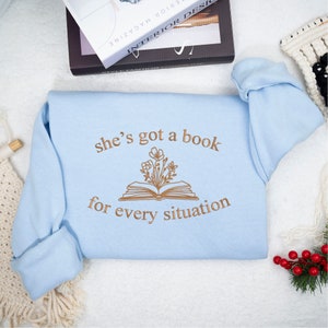 May include: A light blue sweatshirt with the text "she's got a book for every situation" and a brown outline of a book with flowers coming out of it.
