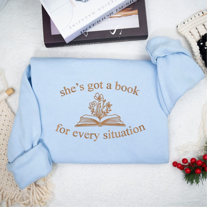 May include: A light blue sweatshirt with the text "she's got a book for every situation" and a brown outline of a book with flowers coming out of it.