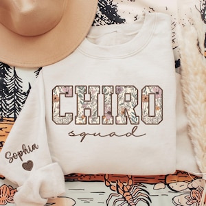 May include: A cream-colored sweatshirt with the text "CHIRo squad" in brown lettering. The letters are outlined in brown and filled with a floral pattern. The word "Sophia" is embroidered on the sleeve with a small heart.