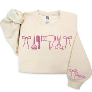 May include: Cream-colored sweatshirt with pink embroidered hair styling tools and bows. The word "Sophia" is embroidered on the sleeve. Features a comb, hair dryer, scissors, and a hair dye brush.