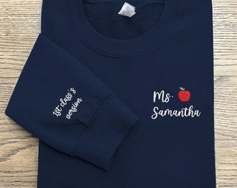 May include: A navy blue sweatshirt with the text "1st class's version" embroidered on the left sleeve and "Ms. Samantha" with a red apple graphic embroidered on the right side.