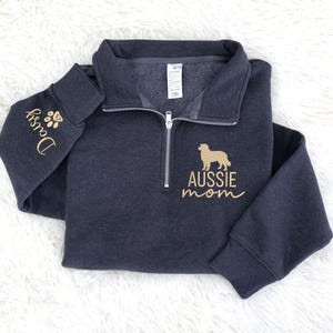 May include: Dark gray quarter-zip sweatshirt with gold embroidery. The left sleeve features the name "Daisy" and paw prints. The chest displays an Australian Shepherd silhouette and the words "AUSSIE mom".