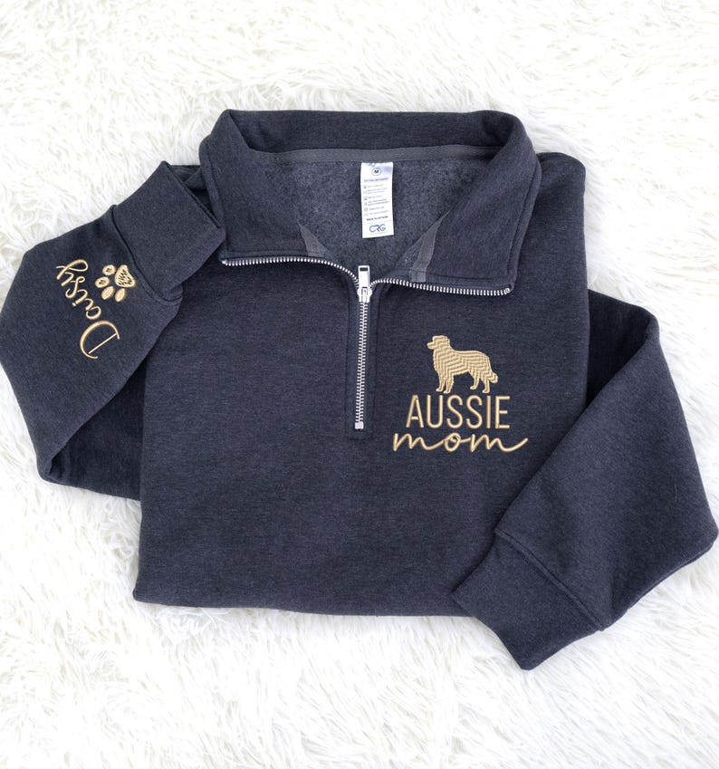 May include: Dark gray quarter-zip sweatshirt with gold embroidery. The left sleeve features the name "Daisy" and paw prints. The chest displays an Australian Shepherd silhouette and the words "AUSSIE mom".