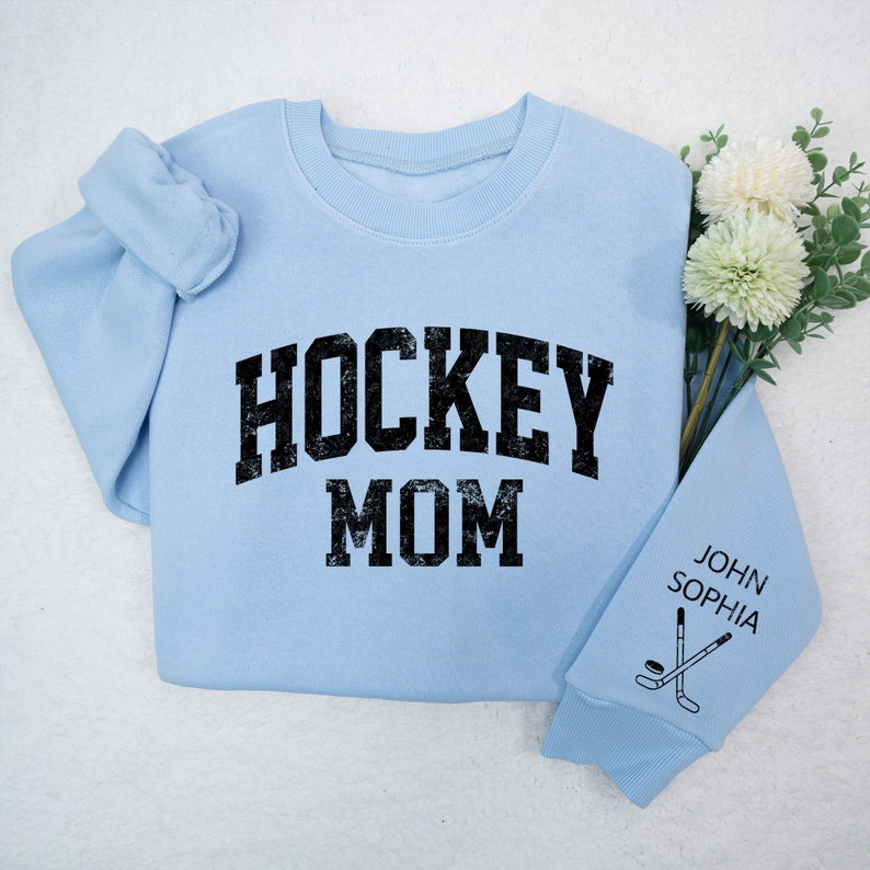 May include: Light blue sweatshirt with the text "HOCKEY MOM" in black. The sleeve has the text "JOHN SOPHIA" and a hockey stick and puck graphic.