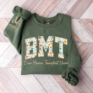 May include: An olive green sweatshirt with the letters "BMT" in a floral pattern. The words "Bone Marrow Transplant Nurse" are printed below the letters. The sleeve has the name "Sophia" embroidered on it.