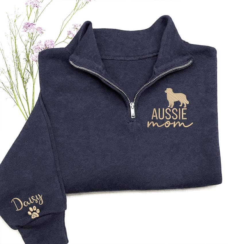 May include: Navy blue quarter-zip sweatshirt with the words "AUSSIE mom" and a gold-colored dog silhouette embroidered on the front. The sleeve has the name "Daisy" and a paw print embroidered in gold.