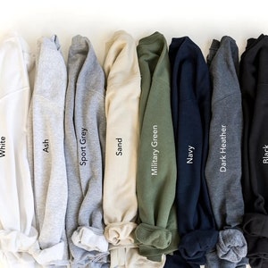 May include: A row of ten folded sweatshirts in various colors, including white, ash, sport grey, sand, military green, navy, dark heather, and black. The colors are labeled below each sweatshirt.