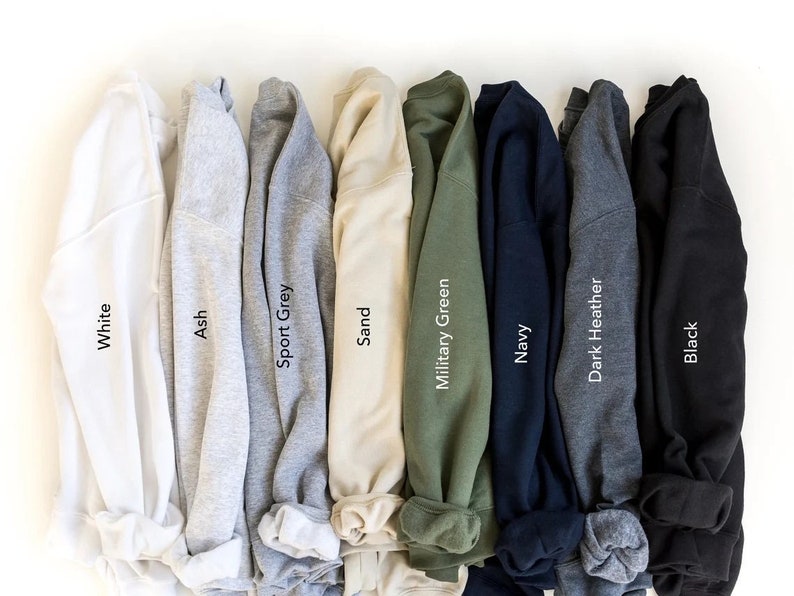 May include: A row of ten folded sweatshirts in various colors, including white, ash, sport grey, sand, military green, navy, dark heather, and black. The colors are labeled below each sweatshirt.